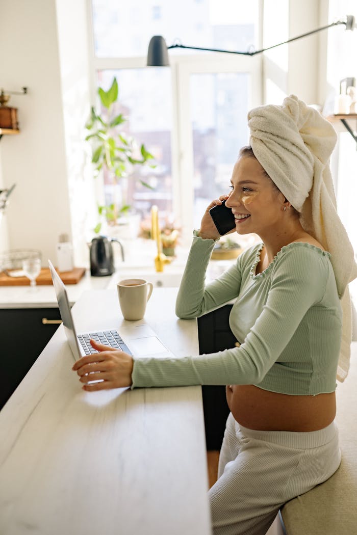 Pregnant woman in kitchen, smiling and multitasking on phone and laptop.